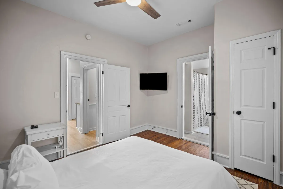 A serene bedroom featuring a neatly made white bed with plush pillows, bathed in soft natural light. A wall-mounted television is visible, and doorways offer a sense of flow into other rooms, hinting at a private, inviting retreat.