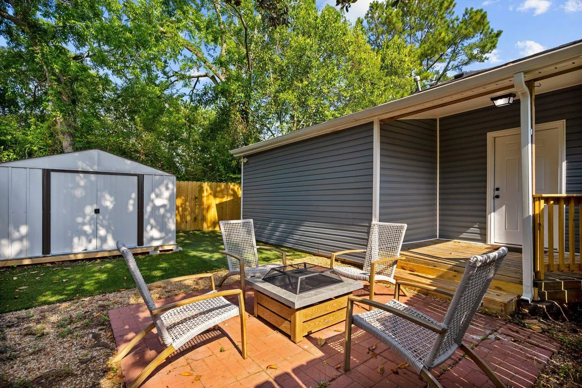 The image displays a welcoming fire pit surrounded by comfortable seating, with the house and garden shed visible in the background. The space is designed for evening conversations, barbecues, and sharing s'mores under the stars.